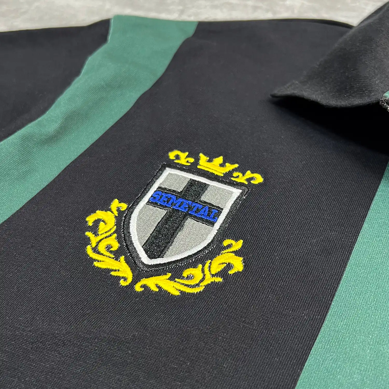 Macro detail of high-density shield crest embroidery on the custom striped polo shirt.